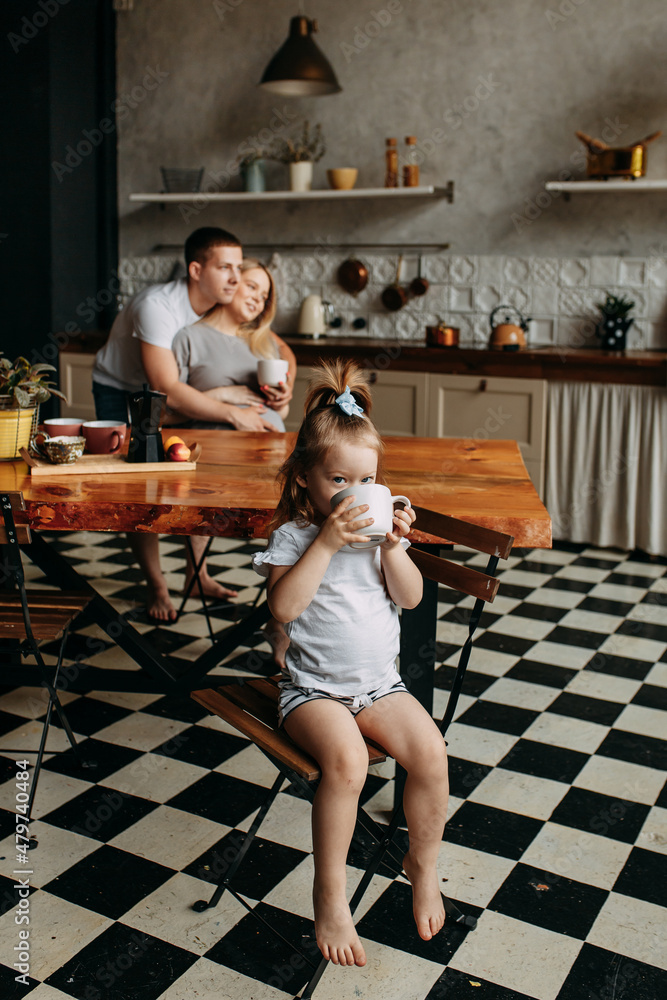 A happy family with one child having breakfast laughing sitting at the dining table in a loft-style kitchen in a cozy house. Pregnant mom dad and little daughter cook together. Selective focus