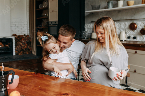 A happy family with one child having breakfast laughing sitting at the dining table in a loft-style kitchen in a cozy house. Pregnant mom dad and little daughter cook together. Selective focus