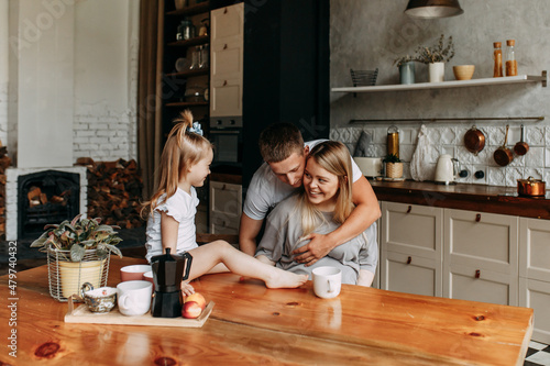 A happy family with one child having breakfast laughing sitting at the dining table in a loft-style kitchen in a cozy house. Pregnant mom dad and little daughter cook together. Selective focus