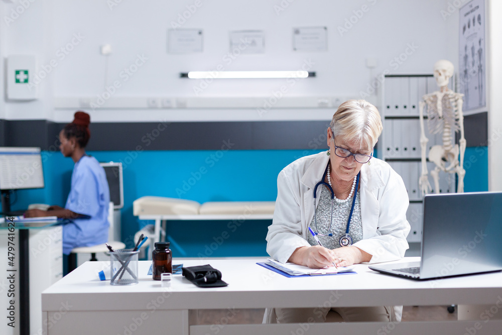 Portrait of senior medic taking notes on checkup papers at desk in office. Woman doctor writing about healthcare, prescription treatment and consultation appointment on clipboard files.