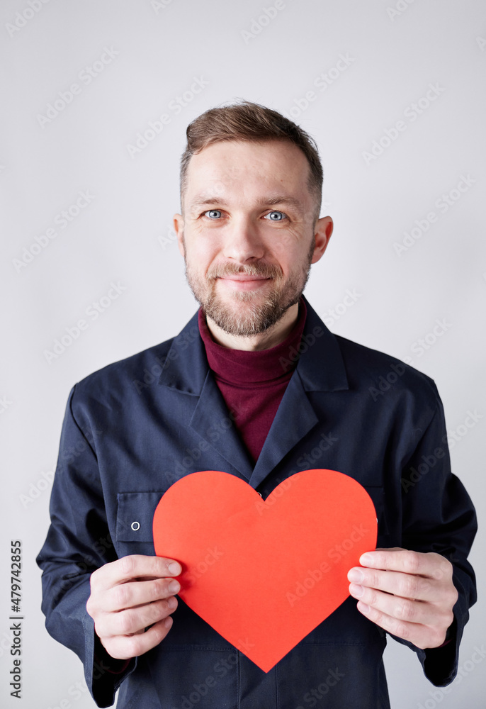 Half length portrait of young bearded hipster man standing by the white wall holding red heart shape. Worker with paper red heart. Romance, relationships concept. Valentine's Day. High quality image Half length portrait of young bearded hipster man standing by the white wall holding red heart shape. Worker with paper red heart. Romance, relationships concept. Valentine's Day. High quality image