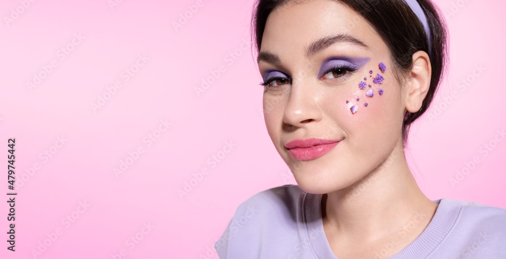 Modern young teen girl with lilac eyeshadow and flowers on healthy smooth cheek skin. Cool woman with lavender make up smiling on pink background. Skincare, make-up cosmetics advertising for youth. Modern young teen girl with lilac eyeshadow and flowers on healthy smooth cheek skin. Cool woman with lavender make up smiling on pink background. Skincare, make-up cosmetics advertising for youth.