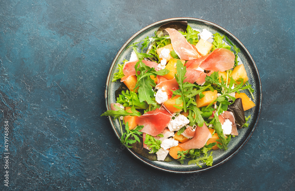 Summer salad with cantaloupe melon, prosciutto, white cheese and arugula on blue table background, top view, copy space