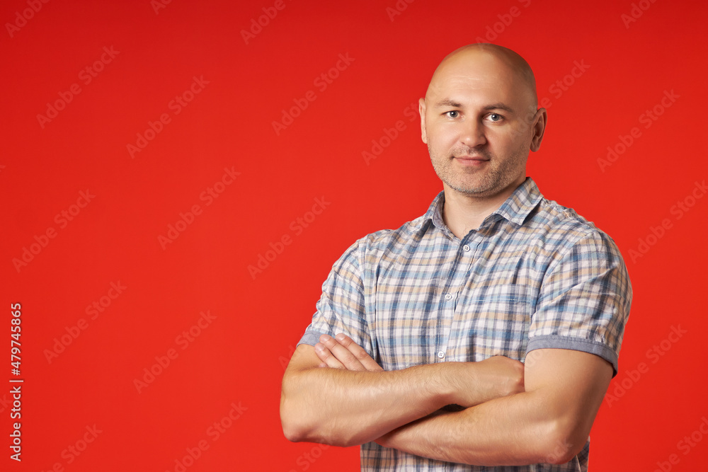 Bald man in a plaid shirt stands on a red background, confidently crossing his arms over his chest. Portrait of a bald guy looking into the camera. Bald man in a plaid shirt stands on a red background, confidently crossing his arms over his chest. Portrait of a bald guy looking into the camera.