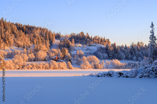 Snow-covered forests and lakes glow in an orange afternoon sunset, by the shores of a winter lake in Norway.