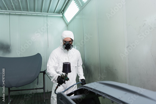 Workman in respirator coloring car part with aerograph in garage.