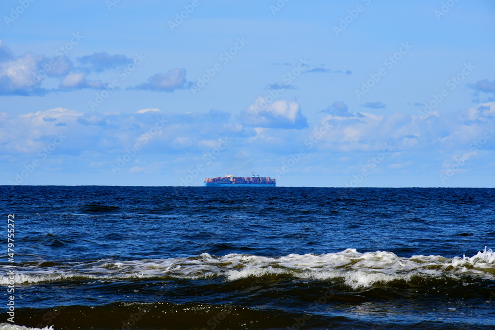 A view on the Polish sea in summer with numerous waves seen from a small sandy beach full of trees and shrubs, as well as with  a big tanker ship seen on the horizon spotted on a cloudy day