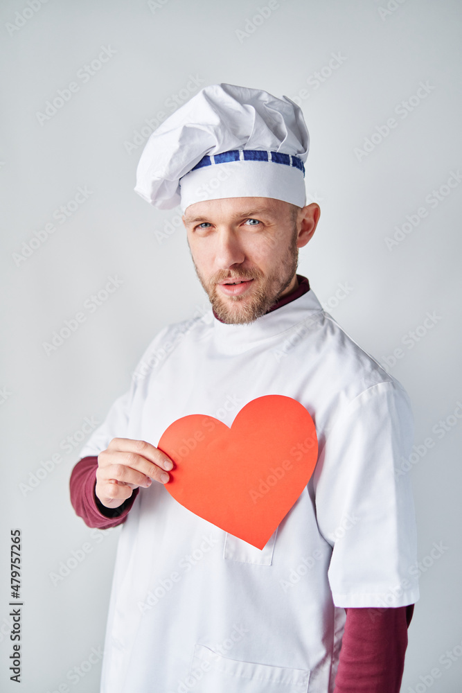 Bearded chef cook or baker man in white uniform with red paper heart as St. Valentine's day concept. Chef cook holding a red heart in front of the workwear. High quality photo
