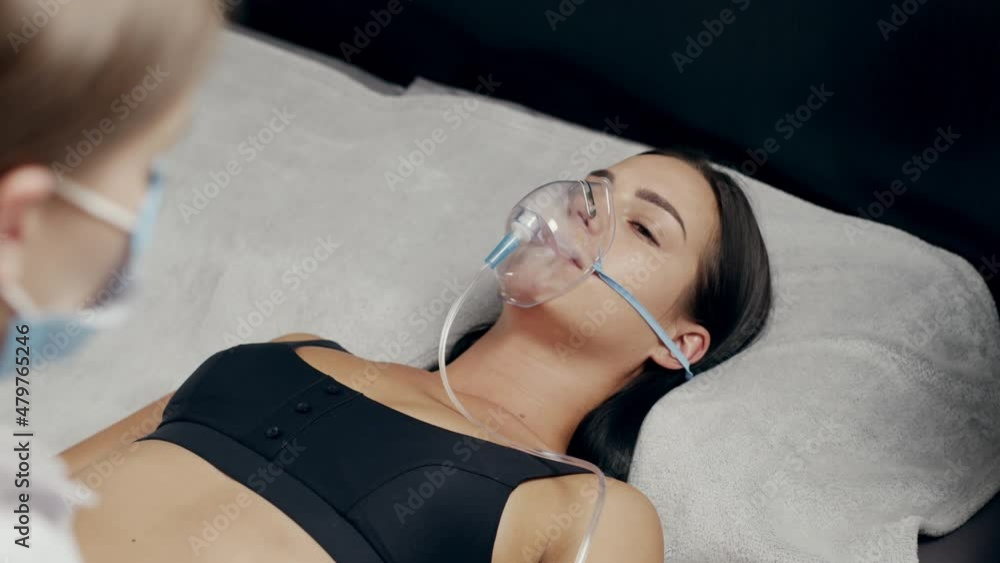 Cinematic 4K. A doctor wearing personal protective equipment or PPE gives an oxygen mask to a Young woman with COVID-19 or coronavirus infection and monitors her health. family doctor medical concept