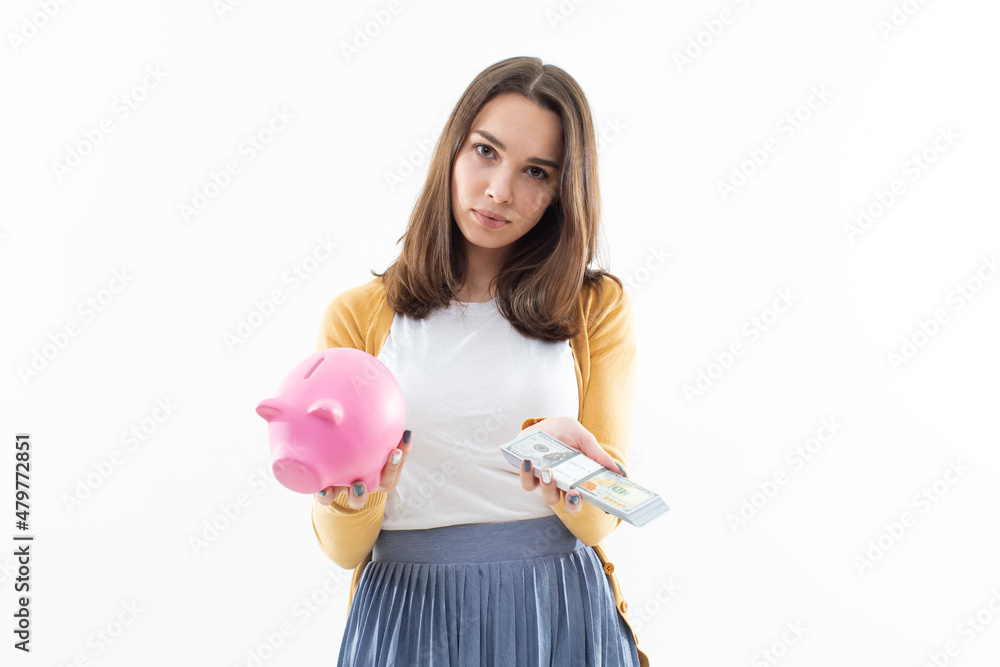 A beautiful brunette holds a pink piggy bank in one hand and a wad of money in the other. Concept of depositing money in a bank A beautiful brunette holds a pink piggy bank in one hand and a wad of money in the other. Concept of depositing money in a bank