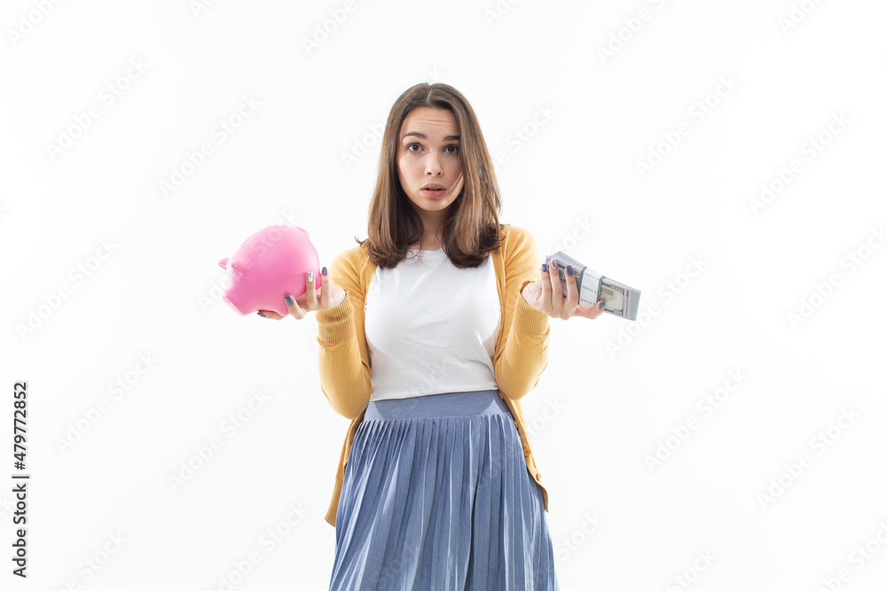 A beautiful brunette holds a pink piggy bank in one hand and a wad of money in the other. Concept of depositing money in a bank A beautiful brunette holds a pink piggy bank in one hand and a wad of money in the other. Concept of depositing money in a bank