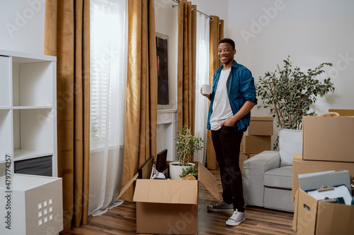 Smiling brunette with dark complexion is standing at window of new apartment, looking out at the view from it, drinking coffee in a mug, living room piled with cardboard boxes with things after move