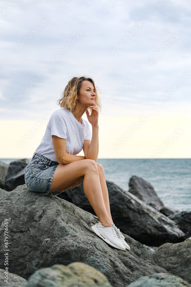 A young attractive Caucasian blonde woman of 35 years sits on the rocks of sea coast. Emerald-colored water, waves splashing on the stones. The woman meditates and relaxes. A young attractive Caucasian blonde woman of 35 years sits on the rocks of sea coast. Emerald-colored water, waves splashing on the stones. The woman meditates and relaxes.