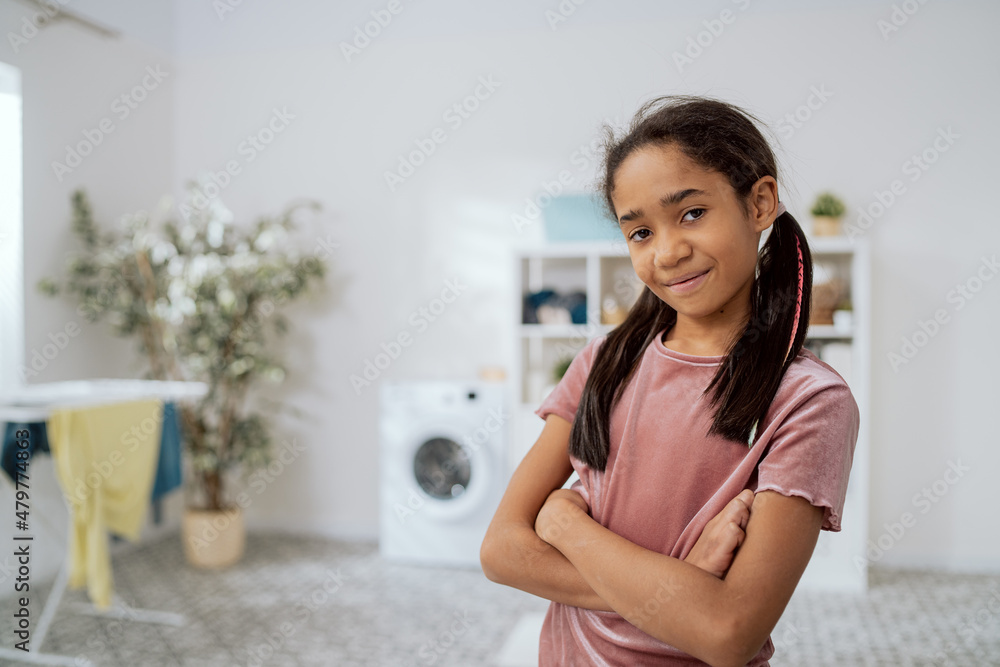 A little girl with dark skin and hair stands in tshirt in middle of laundry room, the bathroom with arms crossed over her chest and a smile, in the background a washing machine, clothes dryer A little girl with dark skin and hair stands in tshirt in middle of laundry room, the bathroom with arms crossed over her chest and a smile, in the background a washing machine, clothes dryer