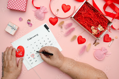 Photography Flat lay on a pink table on which an elderly woman is preparing for the celebration of Valentine's Day marks the date February 14 on the calendar, placed hearts and various paraphernalia on the table