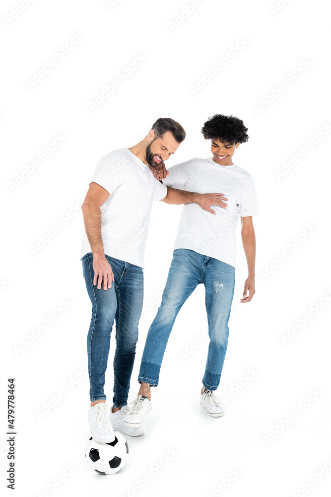 full length view of cheerful multiethnic men in jeans and t-shirts playing football on white.