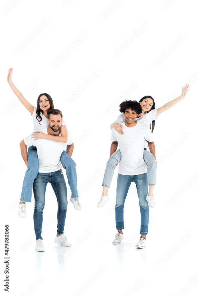 full length view of interracial men piggybacking cheerful women waving hands on white. full length view of interracial men piggybacking cheerful women waving hands on white.