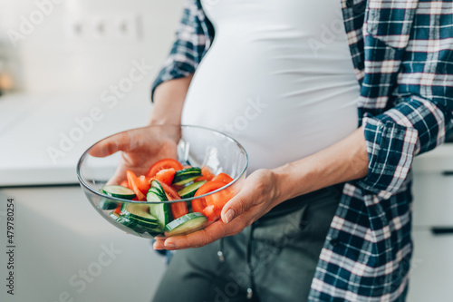 Cropped image pregnant woman hold bowl with vegetable salad of cucumbers and tomatoes in kitchen.
