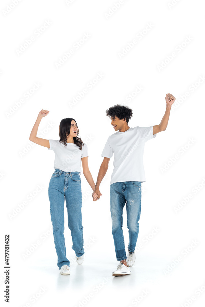 excited multiethnic couple showing win gesture while holding hands and walking on white. excited multiethnic couple showing win gesture while holding hands and walking on white.