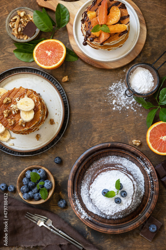 Celebrating Pancake day, healthy breakfast. Delicious homemade american bananas pancakes with blueberries, nuts, caramel, chocolate, red orange on rustic wooden table. Top view flat lay.