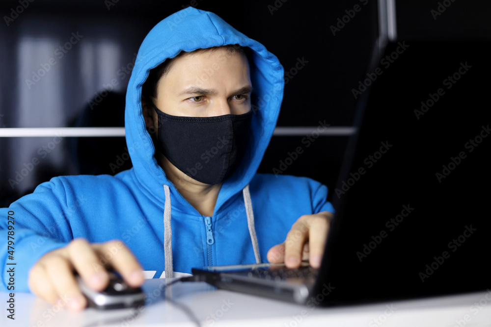 Man in mask and blue hoodie sitting at laptop. Concept of safety office work during coronavirus, cyber crime, hacking and technology Man in mask and blue hoodie sitting at laptop. Concept of safety office work during coronavirus, cyber crime, hacking and technology