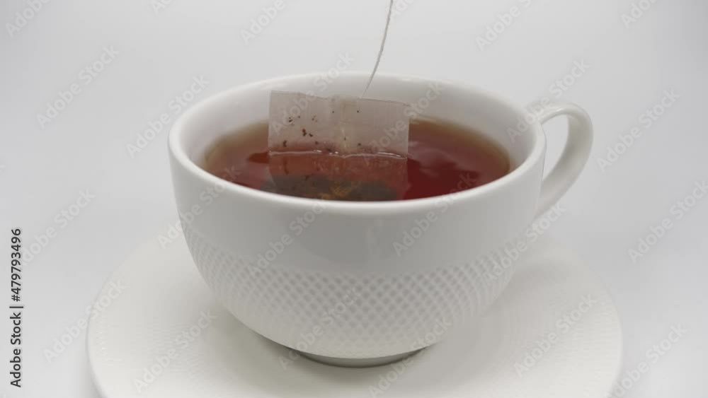 Slow motion. On the table on a white background is a saucer with a cup. The tea bag is brewed in a white cup. Making tea. Hot tea during breakfast. Close-up shooting. Golden drink. Slow motion. On the table on a white background is a saucer with a cup. The tea bag is brewed in a white cup. Making tea. Hot tea during breakfast. Close-up shooting. Golden drink.