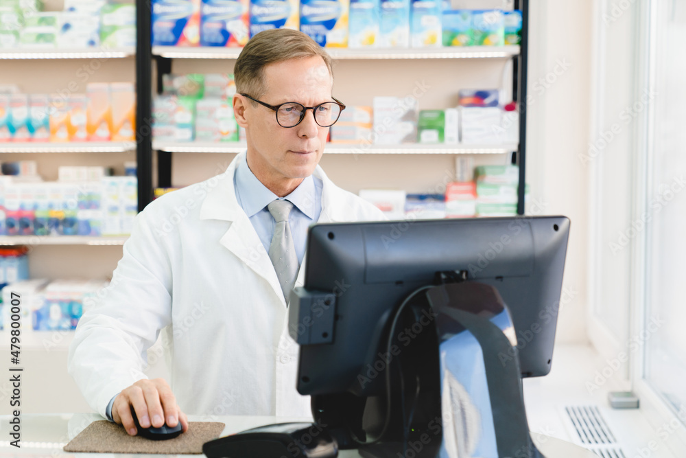 Cash point desk in pharmacy drugstore. Caucasian mature middle-aged chemist pharmacist druggist using computer for checking availability of drugs medicines pills remedy at chemist`s shop