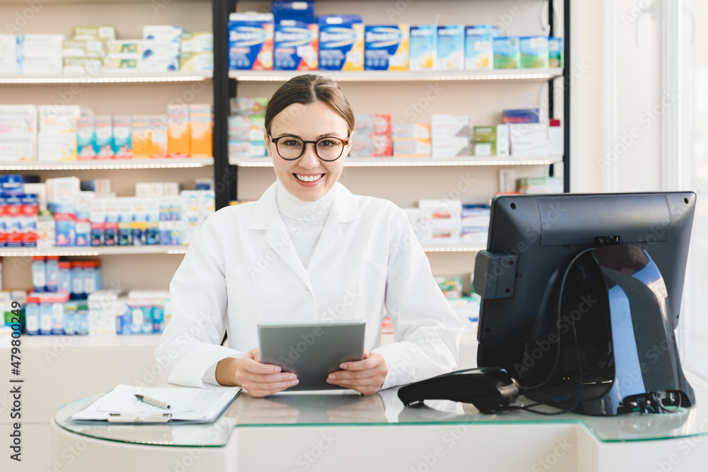 Smiling cheerful caucasian female young pharmacist druggist holding tablet for checking drugs medicines pills remedies on at cash point desk in drugstore pharmacy