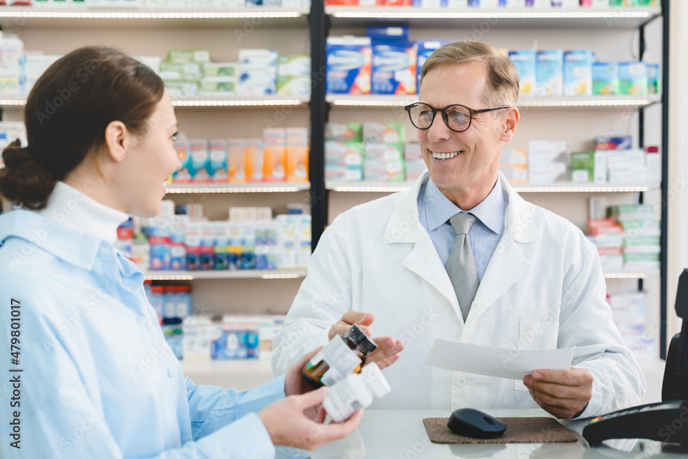 Middle-aged mature male pharmacist druggist advising female customer client buyer remedy, medicines, pills, antibiotics holding prescription in pharmacy