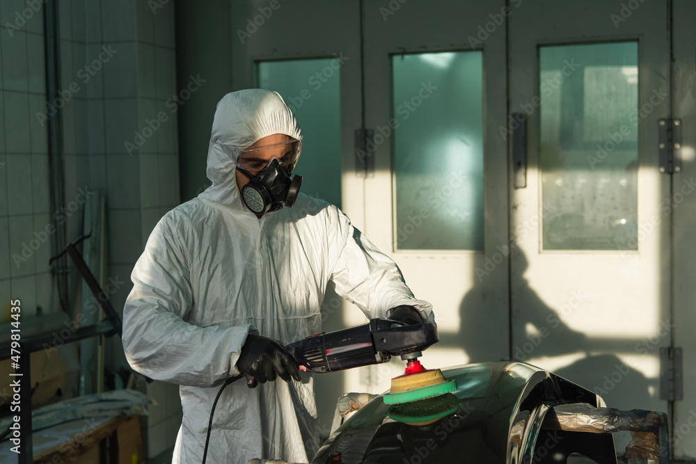 Workman in protective suit and respirator polishing car part in garage. Workman in protective suit and respirator polishing car part in garage.