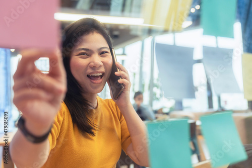 attractive freelance asian female creative person wear casual yellow cloth conversation with smartphone hand point paper note reminder at coworking space new lifestyle with thoughful and freshness 