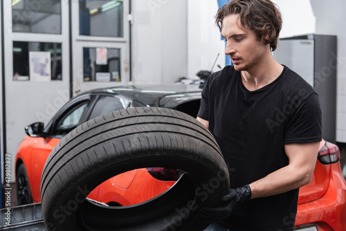 Young workman in gloves holding tire near blurred car in service.