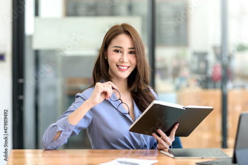 Beautiful young Asian businesswoman sitting holding a book looking at camera with laptop in office. © amnaj