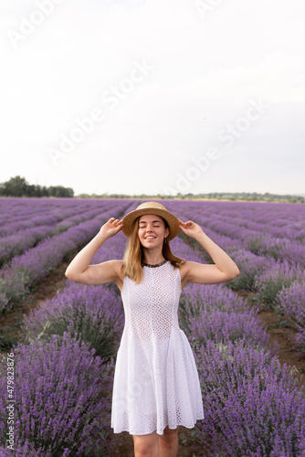 A young beautiful lady stands on a lavender field in provence, dressed in a white dress and a straw hat.