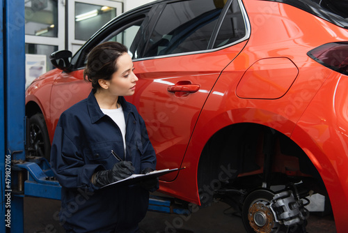 Workwoman in uniform writing on clipboard near car in auto service.