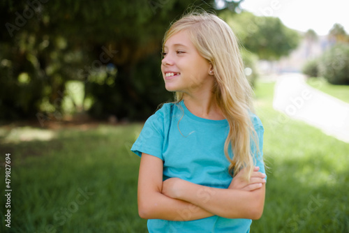 Dreamy rest relaxed Caucasian little kid girl wearing blue T-shirt standing outdoors crossing arms, looks good copyspace