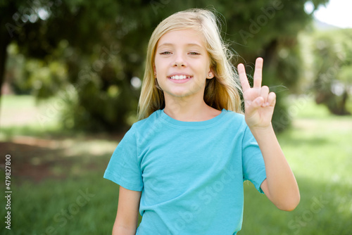 Caucasian little kid girl wearing blue T-shirt standing outdoors showing and pointing up with fingers number two while smiling confident and happy.