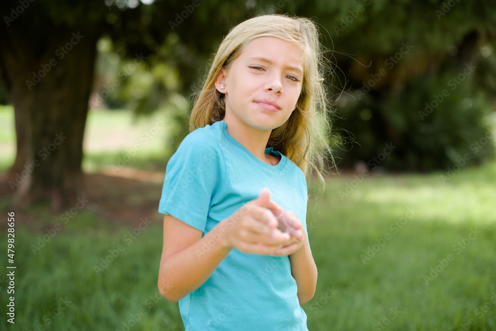 Joyful Caucasian little kid girl wearing blue T-shirt standing outdoors wink and points index fingers at camera, chooses someone, makes finger gun pistol. Joyful Caucasian little kid girl wearing blue T-shirt standing outdoors wink and points index fingers at camera, chooses someone, makes finger gun pistol.