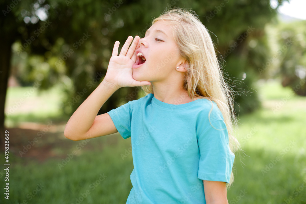 Caucasian little kid girl wearing blue T-shirt standing outdoors shouting and screaming loud to side with hand on mouth. Communication concept.
