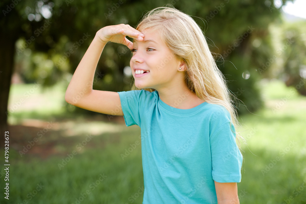 Caucasian little kid girl wearing blue T-shirt standing outdoors very happy and smiling looking far away with hand over head. Searching concept.
