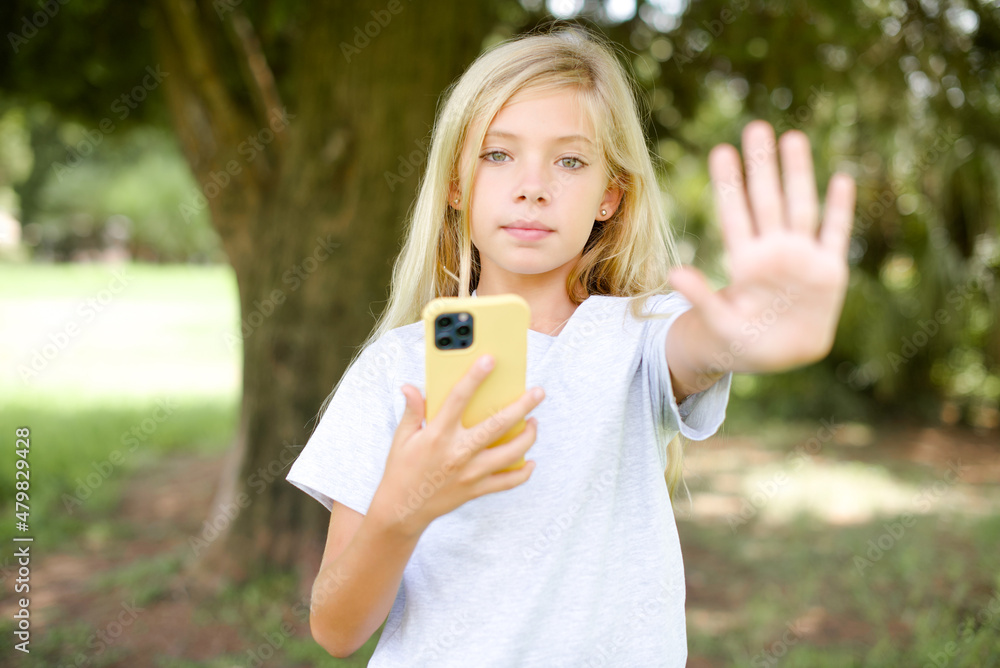 Caucasian little kid girl wearing white T-shirt standing outdoors using and texting with smartphone with open hand doing stop sign with serious and confident expression, defense gesture