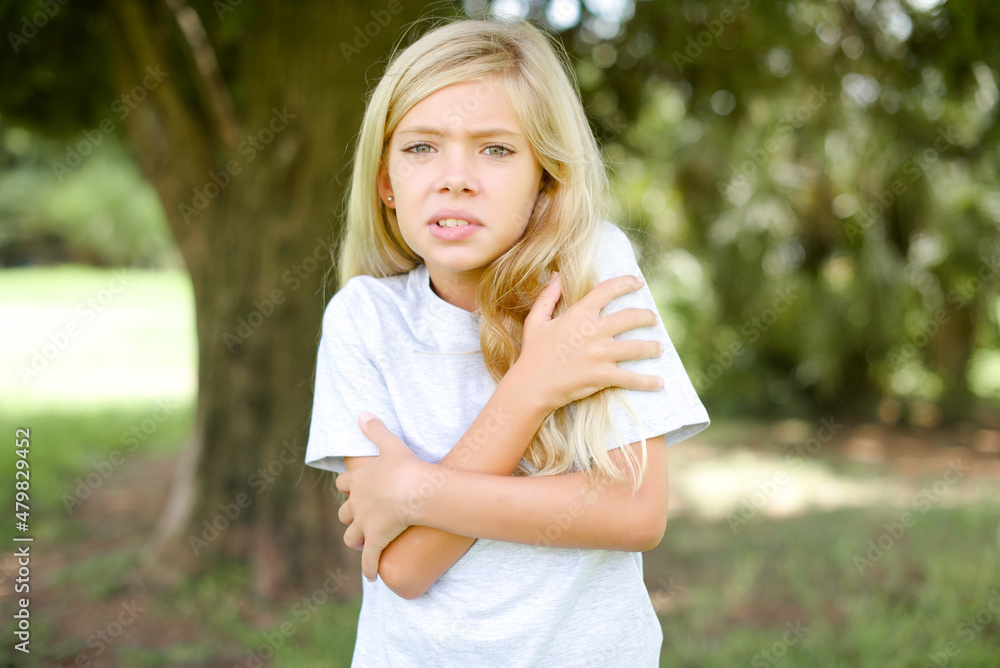 Desperate Caucasian little kid girl wearing white T-shirt standing outdoors trembles and feels cold, hugs oneself to warm up or feels scared notices something terrifying. Desperate Caucasian little kid girl wearing white T-shirt standing outdoors trembles and feels cold, hugs oneself to warm up or feels scared notices something terrifying.