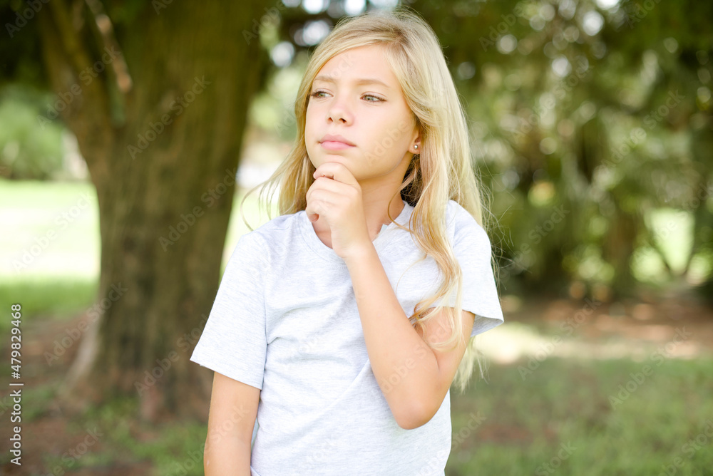 Thoughtful Caucasian little kid girl wearing whiteT-shirt standing outdoors holds chin and looks away pensively makes up great plan Thoughtful Caucasian little kid girl wearing whiteT-shirt standing outdoors holds chin and looks away pensively makes up great plan