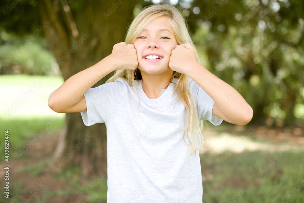 Happy Caucasian little kid girl wearing whiteT-shirt standing outdoors keeps fists on cheeks smiles broadly and has positive expression being in good mood