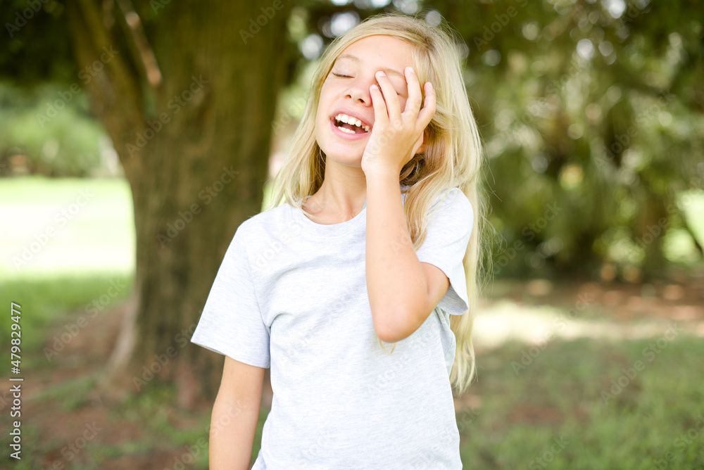 Caucasian little kid girl wearing whiteT-shirt standing outdoors makes face palm and smiles broadly, giggles positively hears funny joke poses