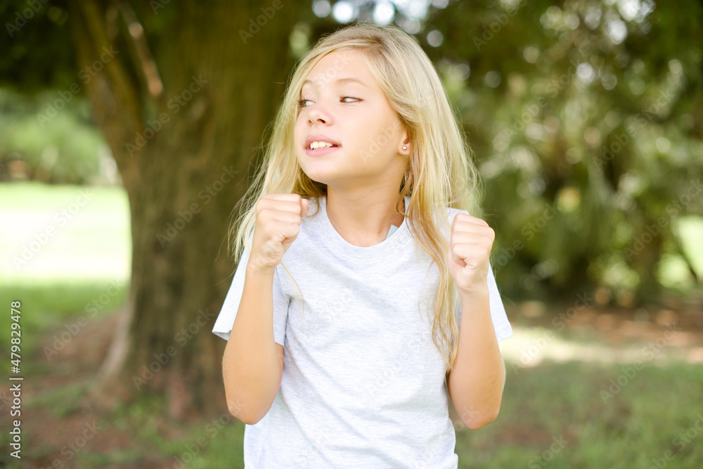 Caucasian little kid girl wearing whiteT-shirt standing outdoors clenches fists and awaits for something nice happened looks away bites lips and waits announcement of results Caucasian little kid girl wearing whiteT-shirt standing outdoors clenches fists and awaits for something nice happened looks away bites lips and waits announcement of results