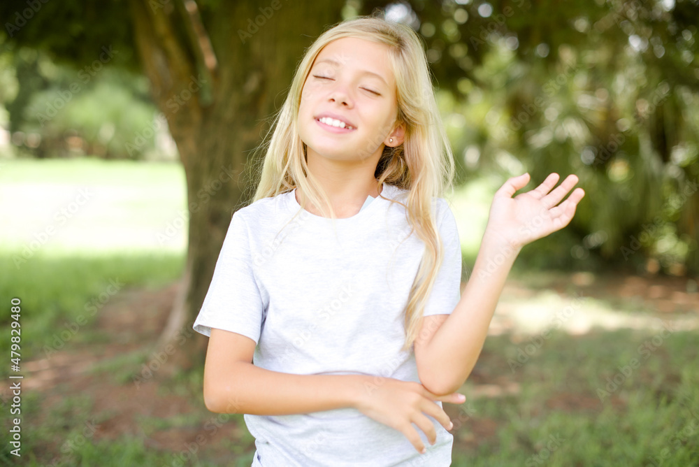 Overjoyed successful Caucasian little kid girl wearing whiteT-shirt standing outdoors raises palm and closes eyes in joy being entertained by friends Overjoyed successful Caucasian little kid girl wearing whiteT-shirt standing outdoors raises palm and closes eyes in joy being entertained by friends