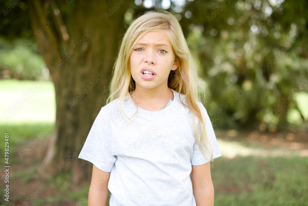 Portrait of dissatisfied Caucasian little kid girl wearing whiteT-shirt standing outdoors smirks face, purses lips and looks with annoyance at camera, discontent hearing something unpleasant