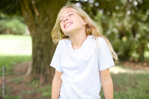 Fotografia Positive Caucasian little kid girl wearing whiteT-shirt standing outdoors with o