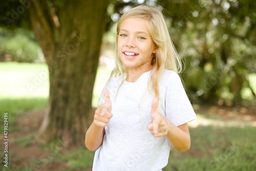 Fototapeta Naklejka Na Ścianę i Meble -  Caucasian little kid girl wearing whiteT-shirt standing outdoors directs fingers at camera selects someone. I recommend you. Best choice